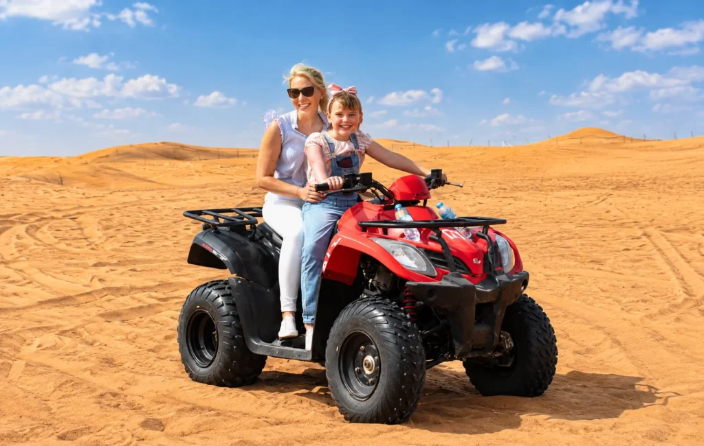 Two tourists experiencing quad biking Dubai on a red ATV across golden sand dunes.