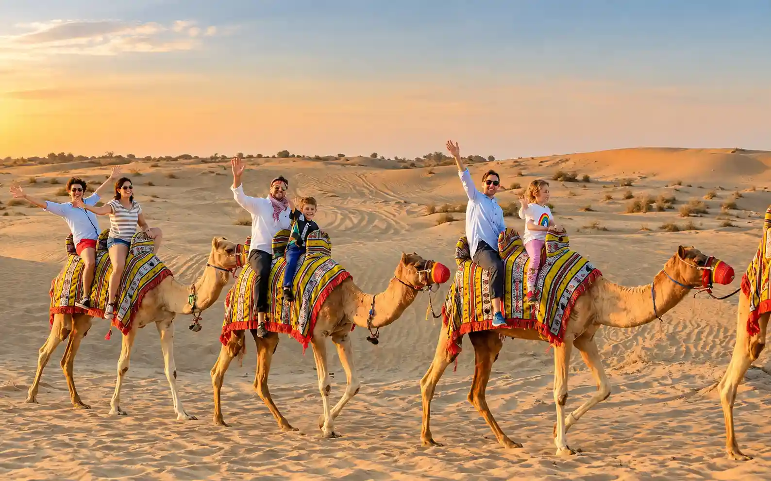 Family riding camels across golden sand dunes during an evening desert safari in Dubai.