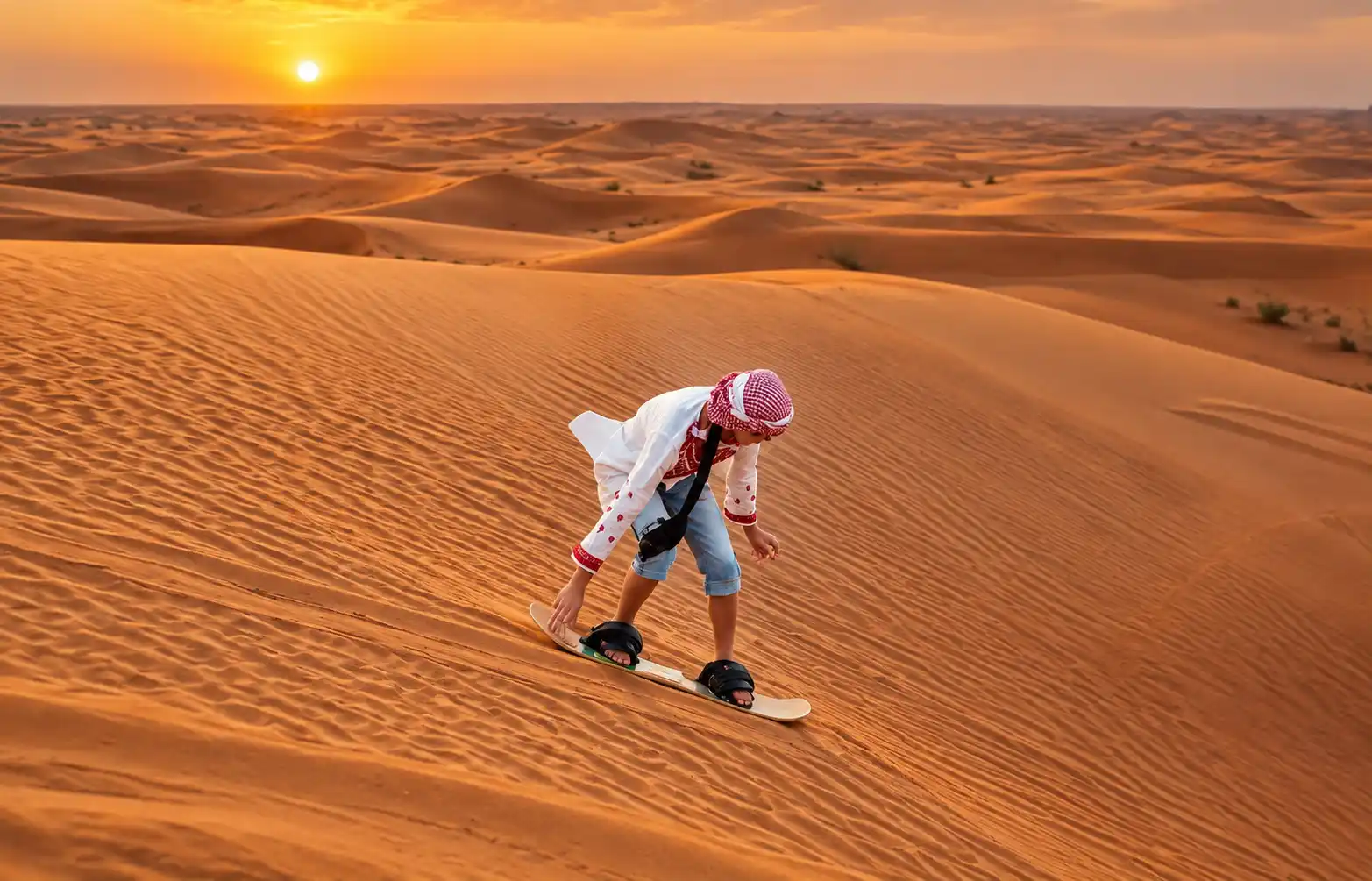 Young boy in traditional headscarf sandboarding down a golden desert dune at sunset