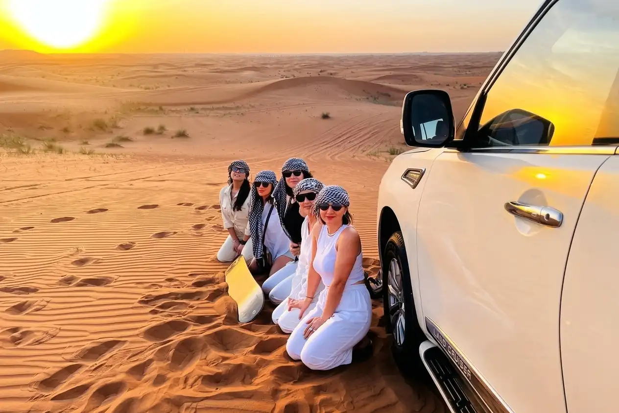 Group of women wearing headscarves sitting on a dune beside a 4x4 at sunset